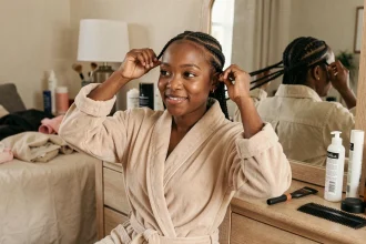 Jeune femme noire réalisant des nattes collées devant son miroir de coiffeuse dans une chambre chaleureuse